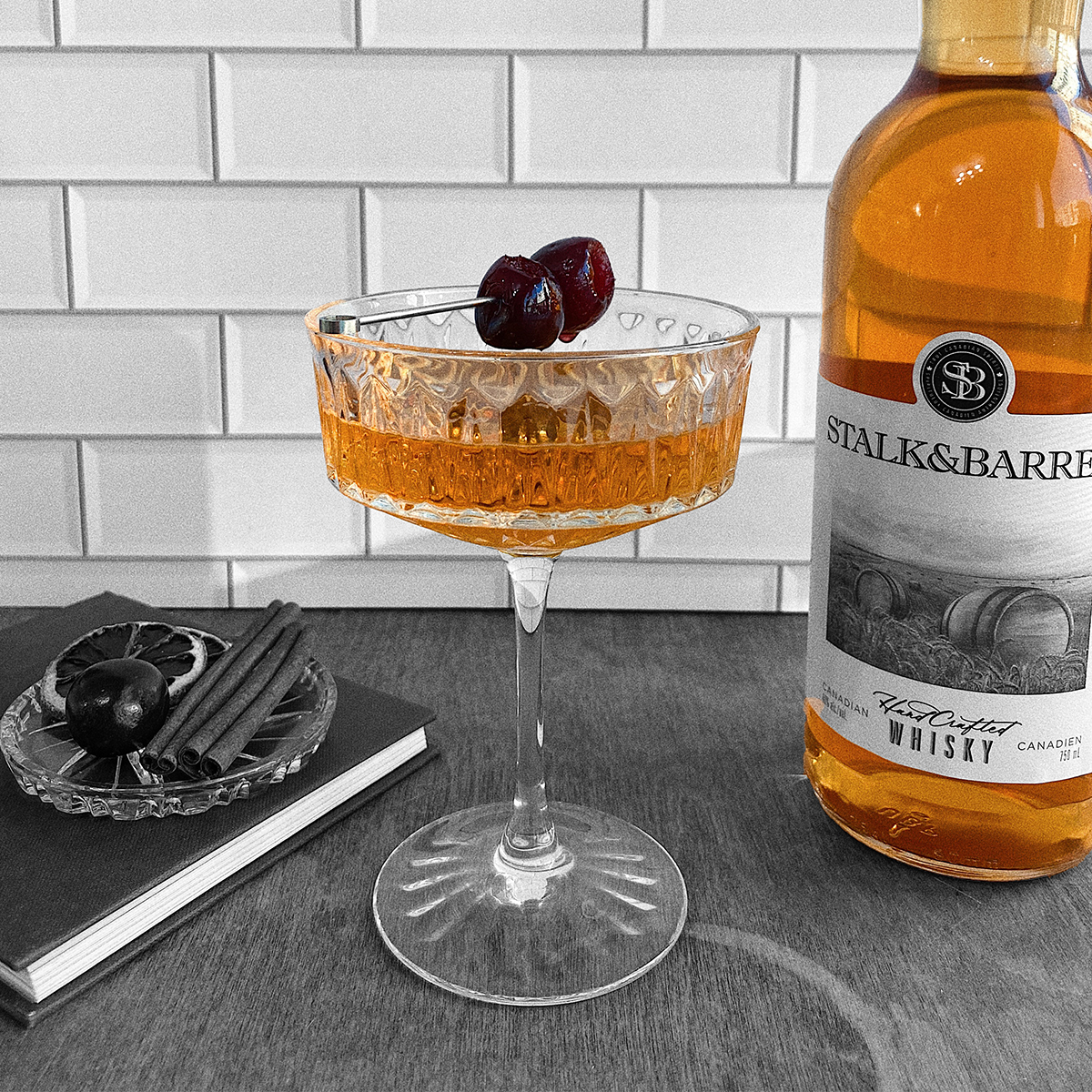 A coupe glass filled with a whisky cocktail, garnisehd with three whisky soaked cherries sits next to a bottle of STALK&BARREL Canadian Whisky